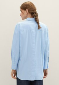Chemise longue en coton bleu clair, dotée d'un empiècement au dos, d'un ourlet arrondi et de poignets boutonnés, avec une texture lisse et une coupe régulière.