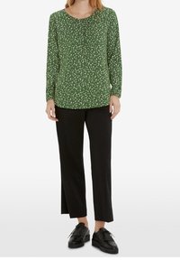 Femme portant un chemisier vert à manches longues à motifs, un pantalon noir droit et des chaussures noires à lacets avec des détails à franges, debout sur fond blanc.