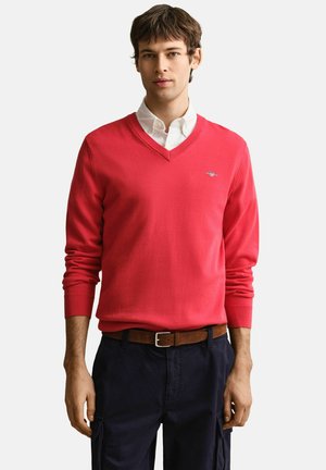 Jonge man met een rode V-hals trui over een wit overhemd, donkere broek met een bruine riem, staand tegen een witte achtergrond.