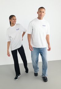 To personer iført oversized hvide t-shirts med "DELIGHT" trykt i blå. Den ene har sorte bukser på, den anden har lyseblå jeans.