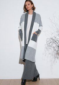 Cardigan lungo grigio e bianco a righe con apertura frontale, silhouette morbida, tasche, abbinato a pantaloni grigi e stivali neri con tacco.