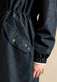 Jūras zilas rakstainas mētelis ar elastīgu jostasvietu, savilktiem aproču galiem un pogāmu kabatu ar uzrakstu un metāla akcentu.