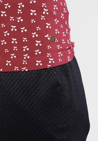 Tissu rouge avec des motifs de cerisiers blancs et une petite étiquette en métal ronde au-dessus de la poche texturée des pantalons noirs.