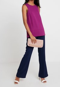 Kobieta w bezrękawnikowej fioletowej bluzce, granatowych szerokich spodniach, w białych szpilkach, trzyma różową kopertówkę na białym tle.