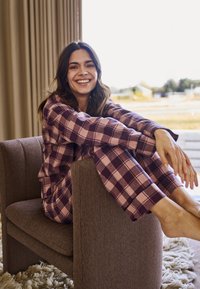 Purple and pink plaid pajama set with long sleeves and pants, worn while sitting on a textured brown chair, featuring a relaxed pose.