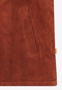 Una tela de ante de color óxido con una textura suave presenta un bolsillo vertical en el lado izquierdo, bordes bien cosidos y una sutil etiqueta de logo.