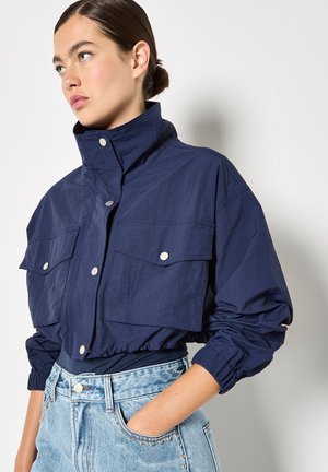 Granatowa krótka kurtka z wysokim kołnierzem, bocznymi kieszeniami i zapięciem na zatrzaski. Posiada elastyczne mankiety i pogniecioną fakturę.