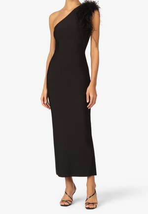 Woman wearing a long black one-shoulder dress with feather detailing on the shoulder and black strappy heels on a white background.