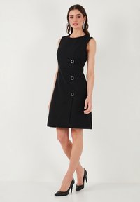 Black sleeveless dress with a fitted silhouette, featuring four square metallic buckles and a knee-length hem. Smooth fabric texture.