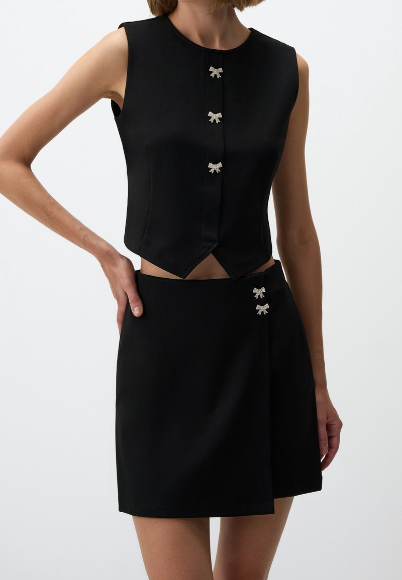 Femme portant un crop top noir sans manches et une jupe assortie, tous deux ornés de petits boutons décoratifs en forme de nœud.