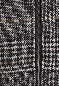 Svart och vitt mönstrat tyg med ett houndstooth-mönster, med en synlig metallblixtlås längs en sida. Strukturerad och vävd look.