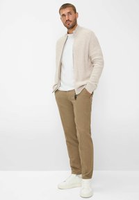 Lichtbeige geribde zip-up trui, wit T-shirt, bruine chinos en witte sneakers. Model staat met handen in de zakken tegen een effen achtergrond.