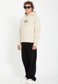 Creme hoodie met zwart grafisch logo, kangoeroezak, en een capuchon met koord; gecombineerd met zwarte wijde broek en witte sneakers. Zwarte zonnebril gedragen.