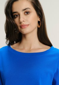 Młoda kobieta z długimi ciemnymi włosami, nosząca złote kolczyki w kształcie kół oraz jasnoniebieską bluzkę, patrzy nieco w stronę kamery z neutralnym wyrazem twarzy.