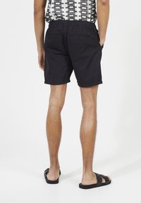 Czarne bawełniane szorty z elastycznym pasem i podwiniętym brzegiem. Dwie tylne kieszenie widoczne, zestawione z czarnymi sandałami.