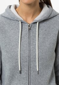 Sudadera gris con cremallera, que cuenta con un forro suave y cordones blancos con puntas metálicas. La tela tiene una textura suave y un diseño casual.