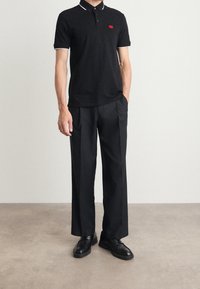 Svart polo shirt med vit kant vid kragen och ärmarna, röd logotyp på bröstet, ihop med lösa svarta byxor och svarta skor.