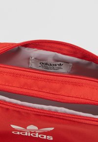 Sacchetto in tessuto rosso con logo Adidas bianco e tre strisce, dotato di chiusura a zip e interno foderato. Etichetta visibile all'interno.
