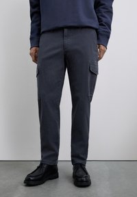 Hombre vestido con pantalones cargo gris oscuro con bolsillos con solapa y zapatos negros de cordones, de pie sobre un suelo gris delante de una pared lisa.