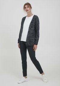 Gilet tricoté bleu foncé avec un motif texturé, au design ouvert à l'avant et aux manches longues, porté sur un t-shirt blanc et associé à un pantalon sombre.