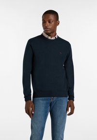 Marineblauer Strickpullover mit Rundhalsausschnitt, gerippten Bündchen und Saum, mit einem kleinen roten Logo auf der Brust, getragen über einem Hemd mit Kragen.