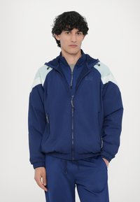 Navy blue windbreaker jacket with light blue accents, zip-up front, adjustable hood, and elastic cuffs, worn over matching pants.