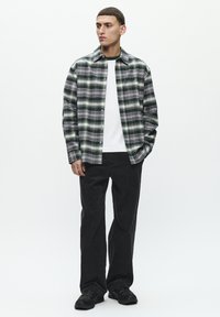 Green and grey plaid shirt with a button front, layered over a white t-shirt; paired with black corduroy pants and black sneakers.