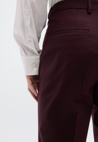 Μπορντό παντελόνια με λεία υφή και εφαρμοστή σιλουέτα, σε συνδυασμό με ένα λευκό πουκάμισο με κουμπιά, που διαθέτει λεπτομέρειες στα μανίκια και λεπτό καρπό.