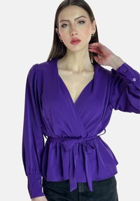 Blouse violette à manches longues, décolleté en V et accent de nœud sur le côté. Tissu lisse avec un aspect légèrement brillant. Assortie à un jean foncé.