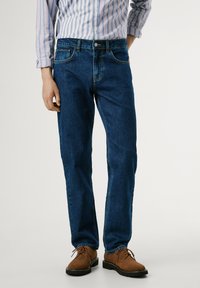 Jeans de mezclilla en azul oscuro con un corte recto, cinco bolsillos y costuras en contraste; combinados con zapatos de ante marrones con cordones.