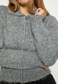 Pilkas megztas kardiganas su visu užtrauktuku, ribinėmis rankogalių ir apačios dalimis, minkšta tekstūra, laisvo silueto, turi apvalų apykaklę ir subtilų raštą visame paviršiuje.