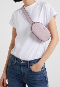 Light pink crossbody bag with quilted texture, oval shape, silver hardware, adjustable strap, worn over a white T-shirt and denim jeans.