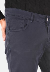 Primo piano di una persona che indossa pantaloni blu scuro con tasche visibili e un maglione nero a maglia cable-knit, con la mano appoggiata sull'anca.