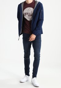 Man med en marinblå zip-up-jacka, vinröd grafisk T-shirt, mörka skinny jeans med reva vid knäet och vita sneakers framför en vit bakgrund.