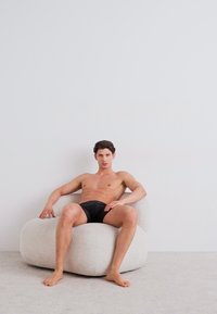 Un fauteuil poire en peluche blanc avec une forme arrondie, présentant une surface texturée. Une personne est assise de manière décontractée, portant un short noir, dans un cadre minimaliste.