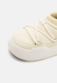 Sneaker color crema con tomaia in tessuto testurizzato, lacci elastici bianchi e suola bianca chunky con branding in rilievo.