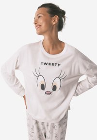 Biała koszula z długim rękawem z dużą grafiką postaci "Tweety". Miękka bawełniana tkanina o luźnym kroju. Szare spodnie piżamowe z nadrukowanymi twarzami z bajek.