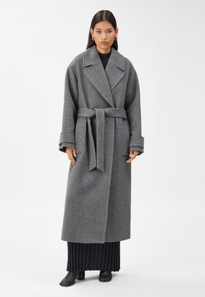 Manteau en laine gris, long, avec une taille ceinturée, de larges revers et des manches retroussées, porté sur une robe noire côtelée et des bottines noires.