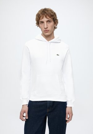 Jeune homme portant un sweat à capuche blanc avec une poche avant et un jean bleu foncé, debout devant un fond clair uni.