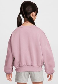 Sudadera oversized rosa clara con dobladillo y puños acanalados, textura suave y hombros caídos. Llevada con pantalones cortos grises.