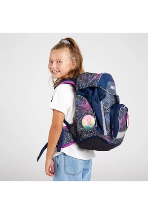 Mädchen mit Pferdeschwanz, lächelnd, trägt ein weißes Hemd und Jeans, mit einem großen dunkelblauen und lila gesprenkelten Rucksack mit bunten runden Aufnähern.