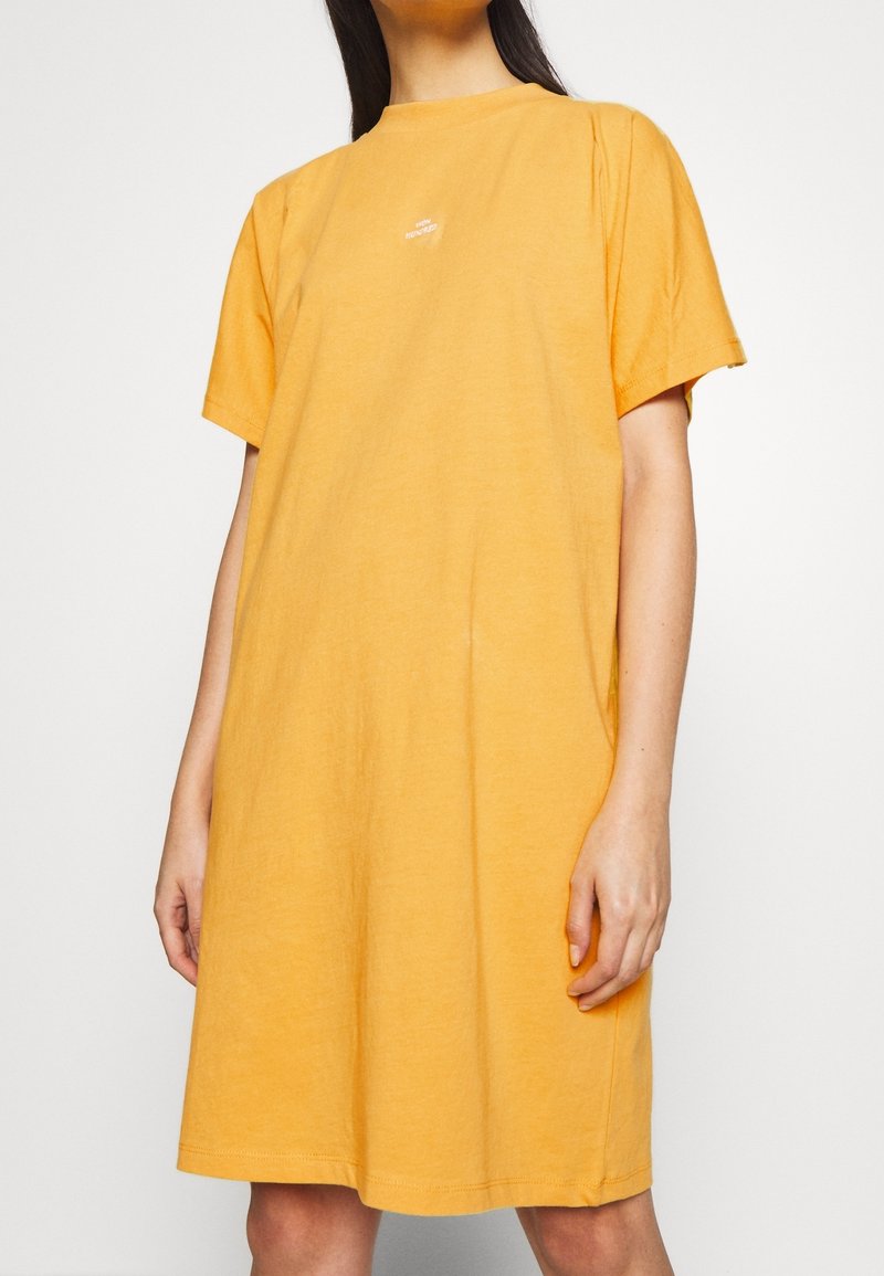 Femme portant une robe jaune ample, manches courtes, longueur au genou, avec un petit texte blanc sur la partie haute de la poitrine.