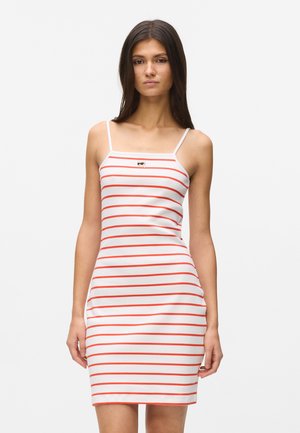 Vrouw die een witte mouwloze jurk draagt met dunne bandjes en horizontale oranje strepen, staand tegen een effen witte achtergrond.