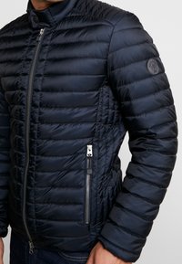 Mörkblå, vadderad puffjacka med ståkrage, blixtlås framtill och två fickor med blixtlås på sidorna. Har logotyp-patch på vänster axel.