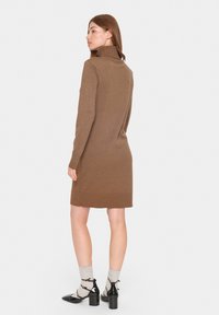 Abito in maglia marrone con colletto alto, texture a coste, forma aderente e orlo che cade sopra il ginocchio; abbinato a calze e scarpe col tacco.