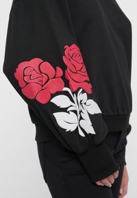 Sudadera negra con grandes gráficos de rosas rojas y acentos florales blancos en la manga izquierda. Textura suave con puños acanalados.