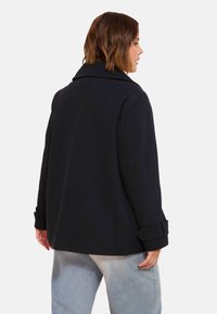 Femme avec des cheveux bruns courts portant un manteau noir avec des poignets à boutons et un jean bleu clair, de profil devant un fond uni.