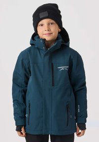 Turkusowa wodoodporna kurtka z zamykanym na suwak kieszonką na piersi, regulowanymi mankietami i logo. Nosi się ją na czarnej koszuli, zestawioną z czarną czapką typu beanie.