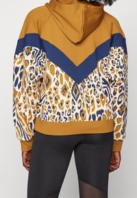 Mikina s kapucí v hnědé, námořnické modré a zvířecím vzoru; má strukturovanou látku, žebrované manžety a volný oversize střih.