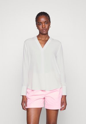 Weiße Bluse mit langen Ärmeln und V-Ausschnitt, lockerer Schnitt und Bündchenärmeln, kombiniert mit pastellrosa Shorts. Glattes Material mit einem dezenten Glanz.